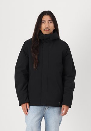 Veste à capuche noire avec fermeture éclair à l'avant, présentant un design élégant, une coupe spacieuse et un tissu texturé. Portée sur un jean bleu clair.