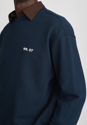 Mann trägt dunkelblauen Pullover mit weißem "NN. 07" Text über braunem Hemd mit Kragen, vor schlichtem hellgrauem Hintergrund.