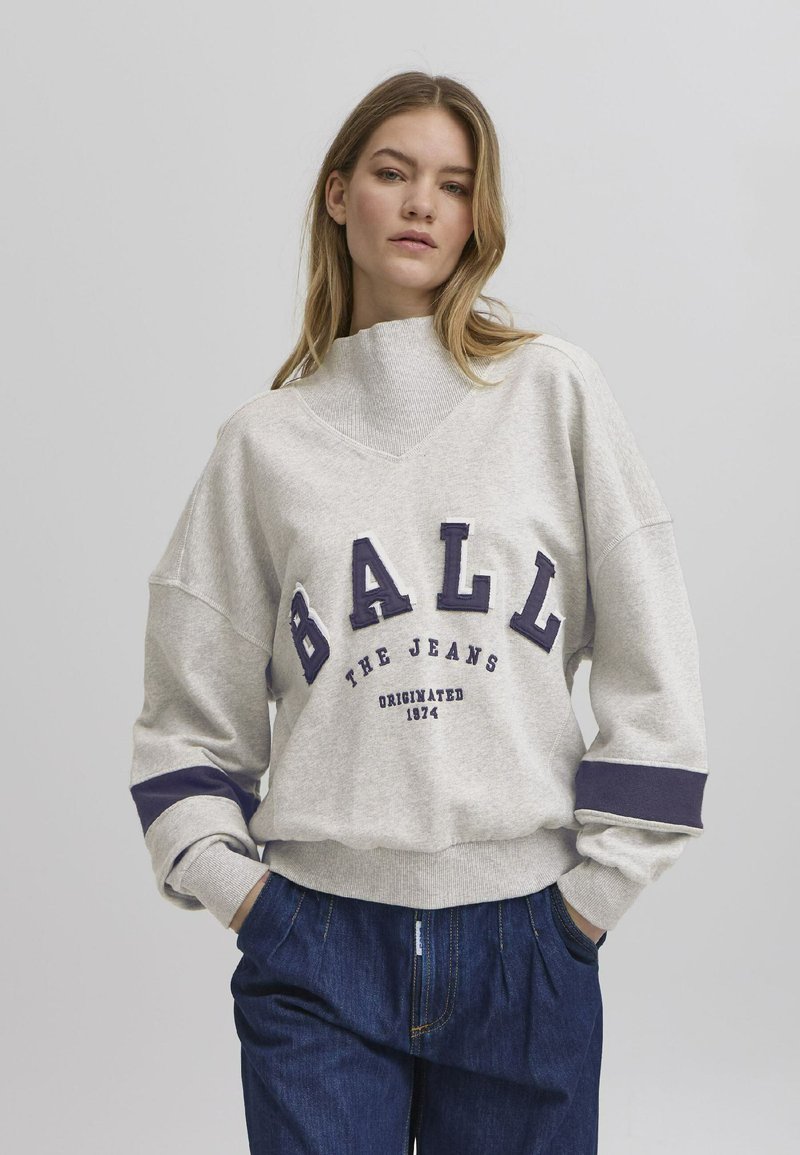 Grå oversized hettegenser med høy krave, som har mørkeblå bokstaver "BALL" og striper. Myk tekstur, avslappet design.