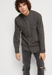 Cămașă din denim gri închis, cu guler cu nasturi, două buzunare pe piept și nasturi din metal. Are un croi relaxat și mâneci lungi.