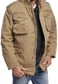 Mężczyzna w beżowej kurtce roboczej z wieloma klapkami kieszeni, częściowo rozpiętej na granatową koszulę i niebieskie dżinsy, trzymający krawędzie kurtki.