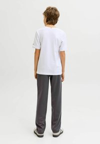 Garçon aux cheveux bruns courts vu de dos, portant un t-shirt blanc, un pantalon gris foncé et des baskets blanches sur un fond clair uni.