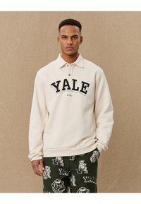 Crème pullover met een polo kraag, met een zwarte "YALE" print en twee zichtbare knopen. In combinatie met donkergroene broek met een patroon.