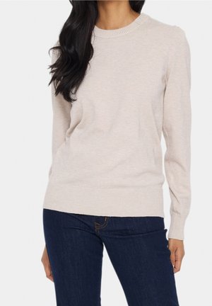 Femme portant un pull en maille beige clair à manches longues avec poignets et ourlet côtelés, associé à un jean bleu foncé sur fond blanc.