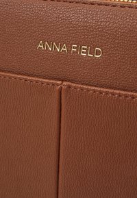 Brun faux læder tegnebogs tekstur med en glat finish, prydet med det guld præget "ANNA FIELD" logo og strukturerede kortlommer.