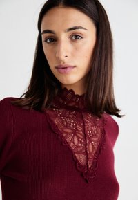 Bordeaux ribbel top met een decoratieve kantkraag met bloemmotieven. Zachte textuur, aansluitend ontwerp en verfijnde kantdetails.