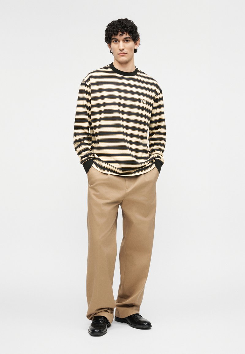 Jonge man die een zwart-beige gestreept shirt met lange mouwen draagt, beige broek met wijde pijpen en zwarte schoenen, staand met handen in de zakken.