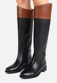 Botas de cuero negras hasta la rodilla con un puño de cuero marrón, cremallera lateral y acentos dorados en las correas. Suela plana y textura suave.