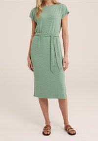 Minttuvihreä mekko, jossa on teksturoitu chevron-kuvio, hihattomat hihat ja solmittava vyö. Suunnitelmassa on suora leikkaus ja sivureiät.