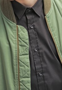 Zielona bomberka z teksturowanym kołnierzem i zapięciem na zamek, noszona na czarnej koszuli z guzikami z klasycznym kołnierzem i okrągłymi guzikami.