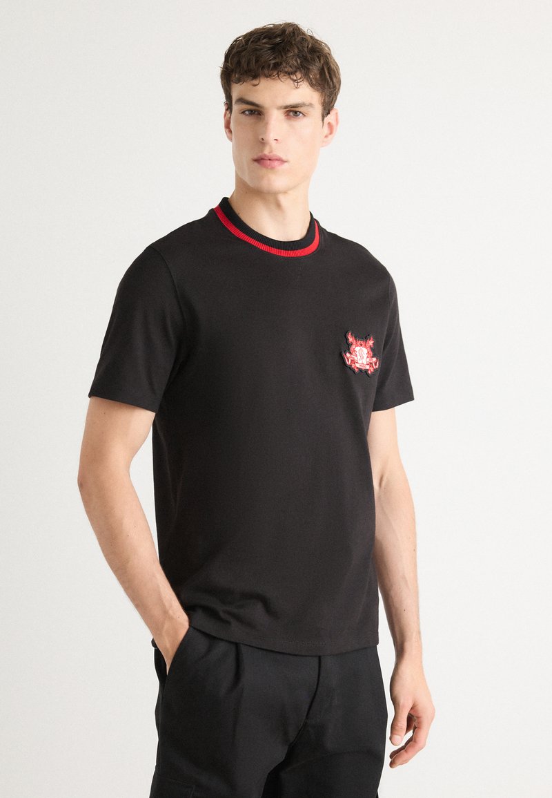 Black cotton t-shirt featuring short sleeves, a round neckline with red trim, and a colorful embroidered design on the chest.
