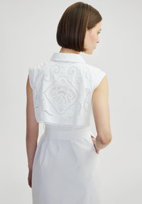 Witte mouwloze blouse met een uitgesneden kant achterkant met ingewikkelde patronen en een kraagontwerp, gecombineerd met een aansluitende witte rok.