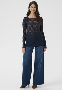 Vrouw in een marineblauw bloemenkant langslurf top, brede blauwe jeans en bruine puntige schoenen, staand tegen een effen witte achtergrond.