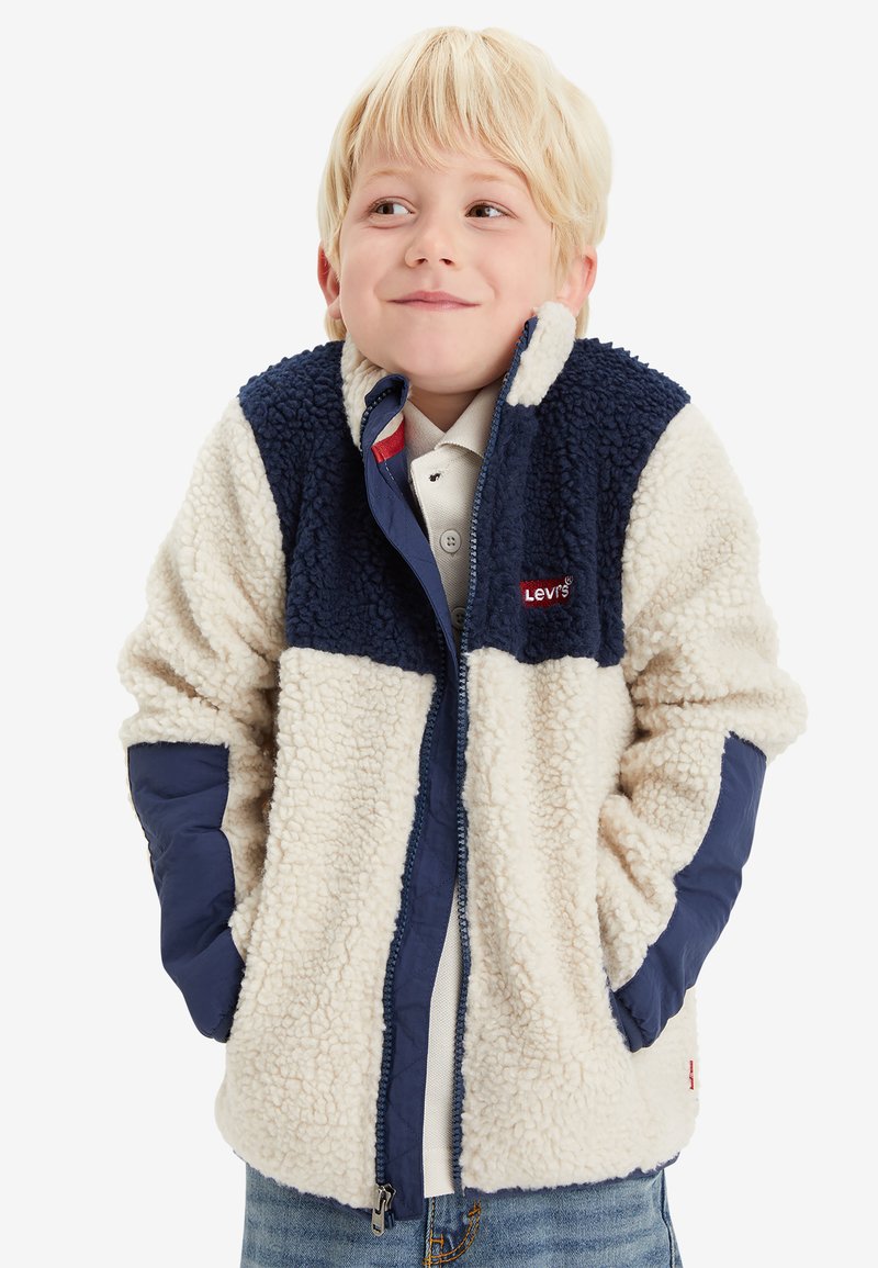 Chaqueta forrada de felpa en crema y azul marino, con cierre de cremallera en el frente, cuello alto y mangas de tela contrastante. Logo rojo en el pecho.