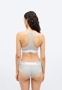 Γυναίκα με μακριά σγουρά μαλλιά που φοράει γκρι αθλητικό τοπ Calvin Klein με σχέδιο racerback και ταιριαστό hipster εσώρουχο, όρθια με την πλάτη στην κάμερα.