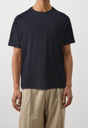 Mand iført en ensfarvet mørkeblå kortærmet t-shirt med en lille lomme og beige løstsiddende bukser.