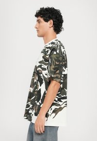 Camo print T-shirt in cotton blend, featuring short sleeves, a crew neck, and shades of green, brown, and white in abstract patterns.
