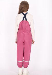 Vaikas su ilgais rudais plaukais vilki rožinius vandeniui atsparius kombinezonus su juodomis petnešomis ir baltomis batų, stovintis iš nugaros ant balto fono.