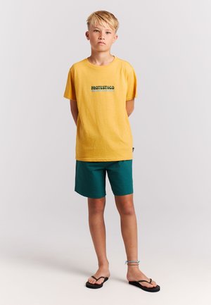 Geel katoenen T-shirt met het logo "PROTEST&CO", gecombineerd met getextureerde groene shorts. Schoeisel: zwarte slippers met een platte zool.