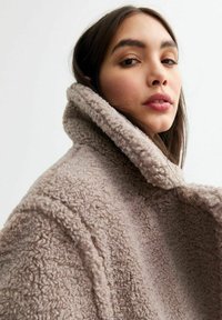 Vezelige beige jas met een hoge kraag, een gestructureerde oppervlakte en brede revers. De stof ziet er zacht en comfortabel uit, met een relaxte pasvorm.