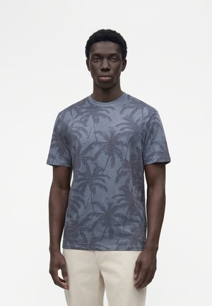 Joven de pie, lleva una camiseta azul con un sutil estampado de palmeras y pantalones beige claro, sobre un fondo liso y claro.