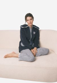 Robe in pile blu navy con rifiniture bianche e dettaglio a forma di cuore. Completo pigiama bianco e blu a quadretti con maniche a volant, indossato seduto su un divano chiaro.