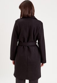 Manteau en laine noir avec des manches longues, taille ceinturée et col à revers. Texture lisse et coupe ajustée, descendant jusqu'à mi-cuisse.