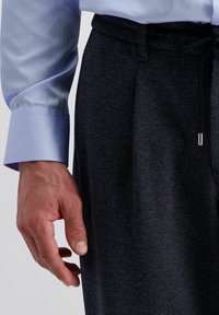 Marineblauwe gemêleerde broek met een trekkoord in de taille, gecombineerd met een lichtblauw satijnen shirt, waarbij een hand nabij het kledingstuk is geplaatst.