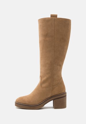 Bota alta de ante en tono marrón claro, con un grueso tacón cuadrado, que presenta adornos de costura y una punta redondeada. Diseño minimalista con una pestaña de tiro en la parte superior.