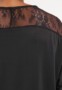 Czarna koszula z przezroczystym panelu koronkowym na plecach z kwiatowym wzorem, kontrastującym z gładkim materiałem głównej części odzieży.