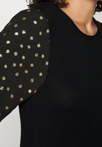 Haut en tricot noir avec manches longues et motif de pois dorés brillants. Le col est rond, le tissu est lisse et ajusté.