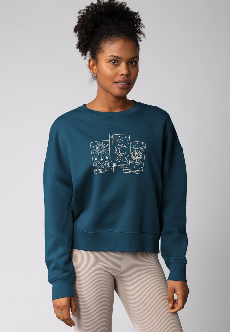 Sweatshirt teal avec une coupe décontractée, présentant un graphisme de cartes de tarot. Manches et ourlet côtelés, texture douce, porté avec des leggings beiges côtelés.