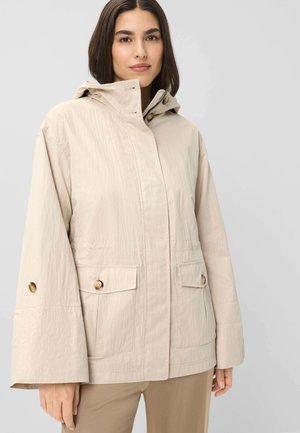 Frau trägt eine beige Kapuzenjacke mit großen geknöpften Taschen und ausgestellten Ärmeln, steht vor einem schlichten hellen Hintergrund.