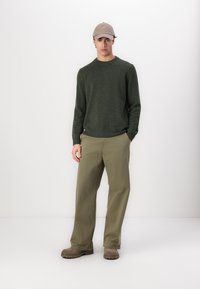 Ung mann iført olivengrønn genser, beige caps, khaki bukser og brune støvler som står mot hvit bakgrunn med hendene i lommene.