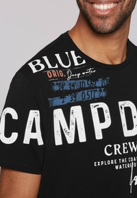 Camiseta de algodón negra con las letras blancas en negrita "CAMP" y "BLUE", con acentos de texto en naranja y azul y diversas coordenadas geográficas impresas.