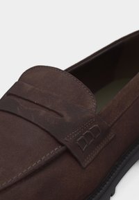 Καφέ μοκασίνι από σουέντ με μύτη μοκασίνου, διακοσμητικό λουράκι στο επάνω μέρος, εμφανή ραφή και μαύρη λαστιχένια σόλα.