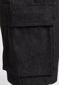 Zwarte denim shorts met een grote, gestructureerde cargozak aan de zijkant, zichtbaar stiksels en een gevouwen zoomdetail.