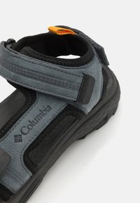 Sandale Columbia grise avec des sangles ajustables, une surface texturée, une semelle en caoutchouc et un onglet d'accent orange. Design léger et sportif.