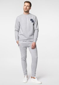Szara bluza z okrągłym dekoltem i granatowym logo na piersi, w zestawie z dopasowanymi szarymi spodniami dresowymi i białymi trampkami.