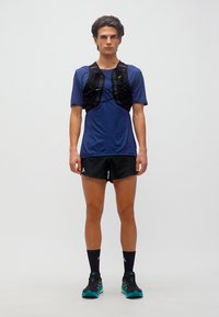 Νεαρός άνδρας που στέκεται, φοράει μπλε αθλητική μπλούζα, μαύρο γιλέκο για τρέξιμο, μαύρο σορτς, μαύρες κάλτσες και μαύρα παπούτσια τρεξίματος με τυρκουάζ σόλες.