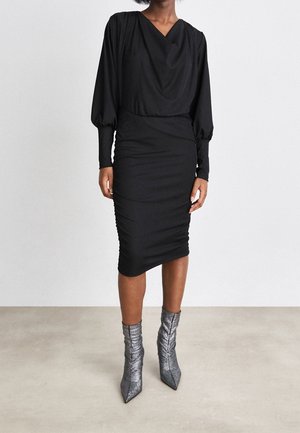 Kobieta w czarnej, marszczonej, midi sukience z długim rękawem i balonowymi rękawami oraz metalicznych, spiczastych botkach do kostki stojąca na neutralnej podłodze.