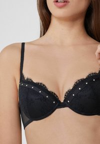 Sostén de encaje negro con bordes festoneados, copas acolchadas y tachuelas plateadas decorativas a lo largo de la banda. Tirantes ajustables y lados de malla transparente.