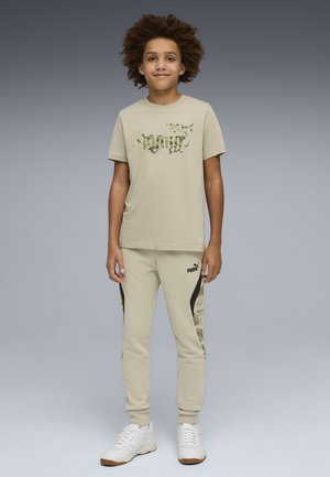 Ragazzo in piedi, indossa una t-shirt beige Puma e pantaloni da jogging coordinati con dettagli mimetici, scarpe da ginnastica bianche, su uno sfondo a gradiente grigio.