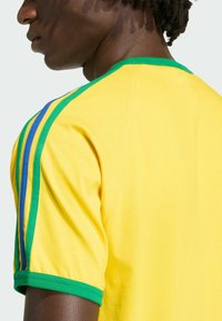 Zbliżenie osoby noszącej żółtą koszulkę z zielonymi wykończeniami i niebiesko-zielonymi paskami na ramieniu, widzianej z boku i tyłu.