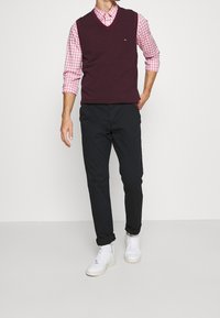 Bordeauxkleurig gebreid vest over een roze geruit overhemd, zwarte broek en witte sneakers. Opvallende details zijn een klein logo op het vest.