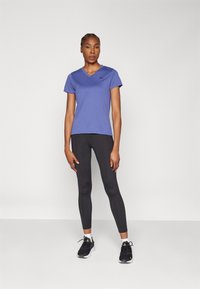 Lilla T-skjorte med kort erme og v-hals, svarte tettsittende leggings, svarte sportssko med hvite detaljer, stående mot en helt hvit bakgrunn.