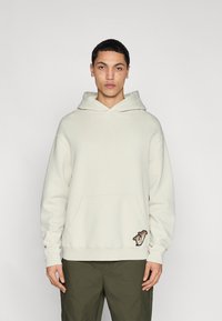 Jeune homme aux cheveux courts et taches de rousseur portant un sweat à capuche crème avec un petit motif près de la poche et un pantalon vert olive, debout devant un fond blanc.