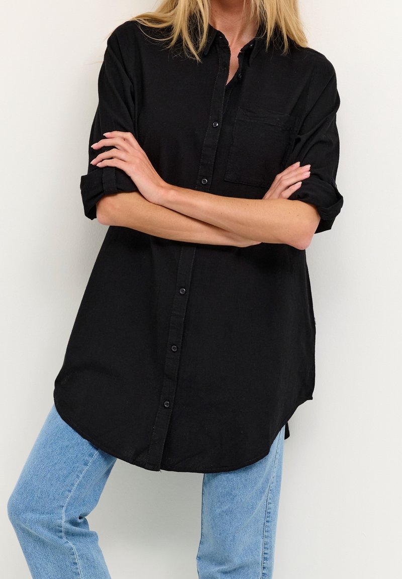 Femme aux bras croisés portant une longue chemise noire boutonnée et un jean bleu clair, sur fond blanc uni.