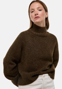 Pull marron en maille avec un col côtelé haut, des manches amples et une texture douce. Porté sur un pantalon blanc, soulignant le contraste des couleurs.