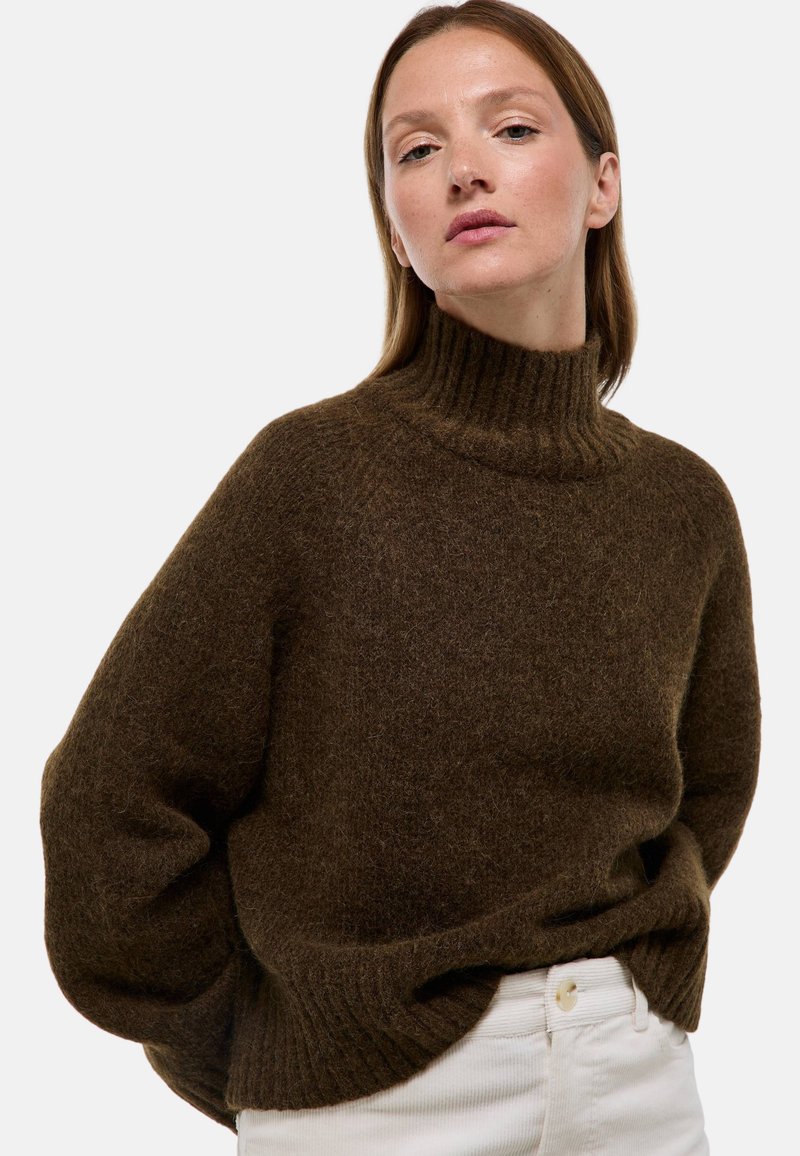 Pull marron en maille avec un col côtelé haut, des manches amples et une texture douce. Porté sur un pantalon blanc, soulignant le contraste des couleurs.